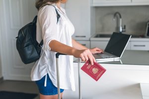 Woman carrying luggage and holding her passport while checking her laptop.